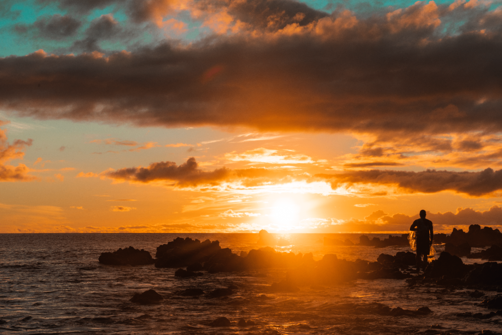 Dramatic orange sunset over the ocean with dark clouds and a lone surfer standing among shoreline rocks