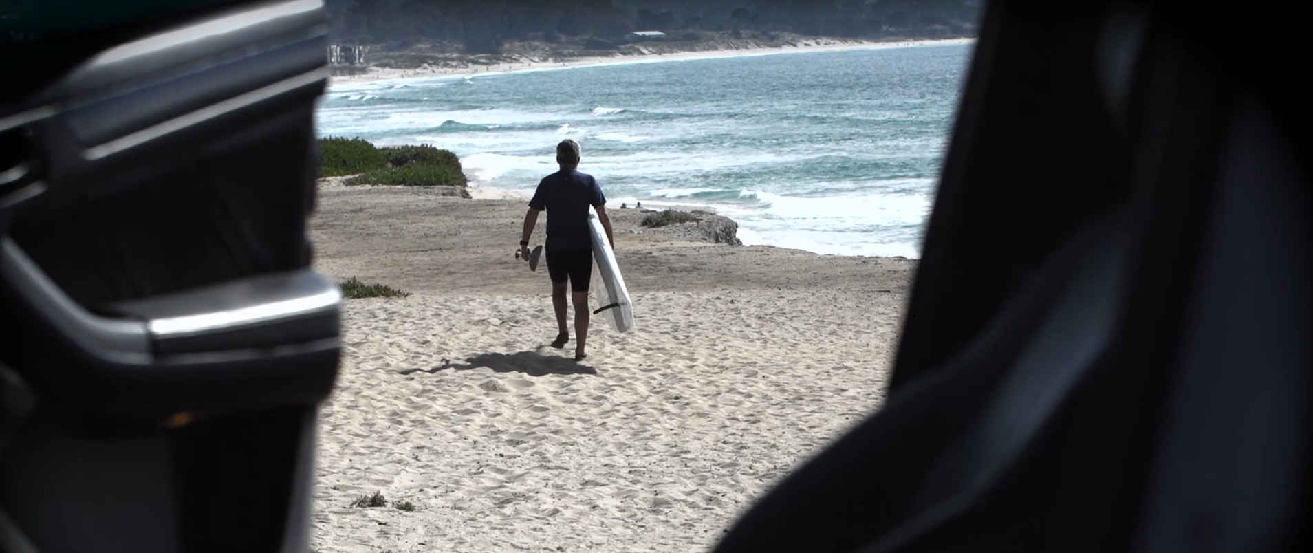 Surfer walking toward the beach with board in hand framed from a vehicle interior