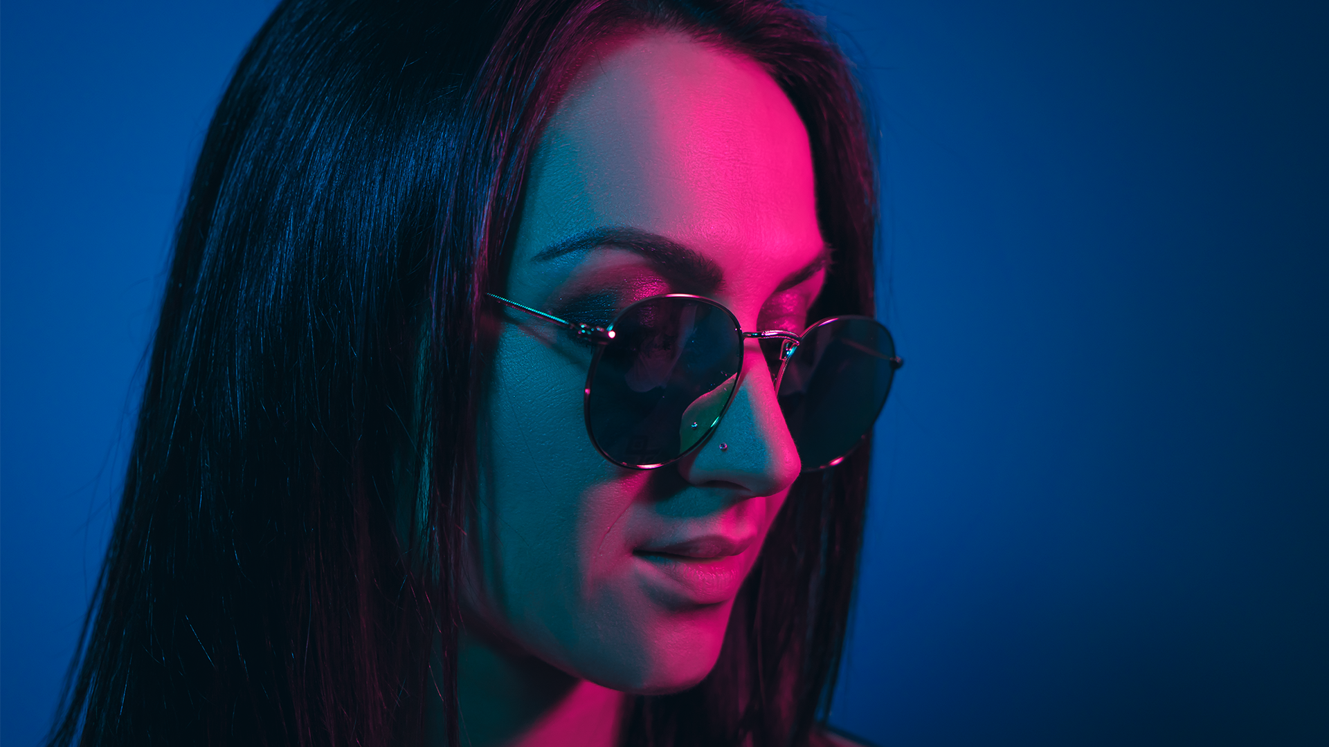 Portrait of a woman in aviator sunglasses lit by magenta and teal neon against a deep blue backdrop