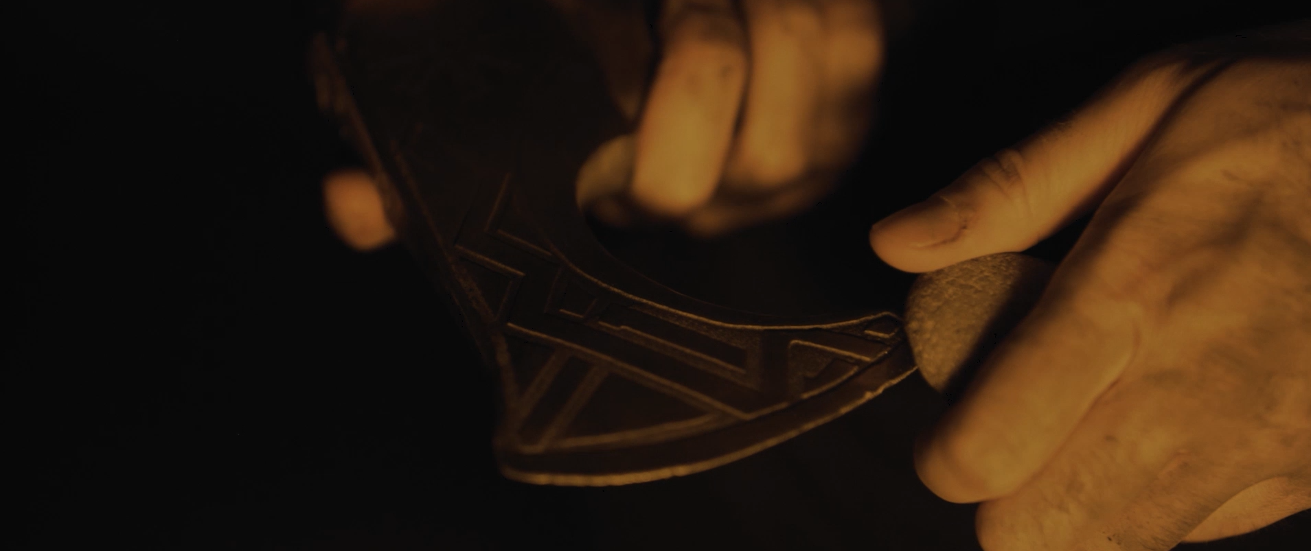 Hands carving an axe head by warm firelight in an intimate close-up