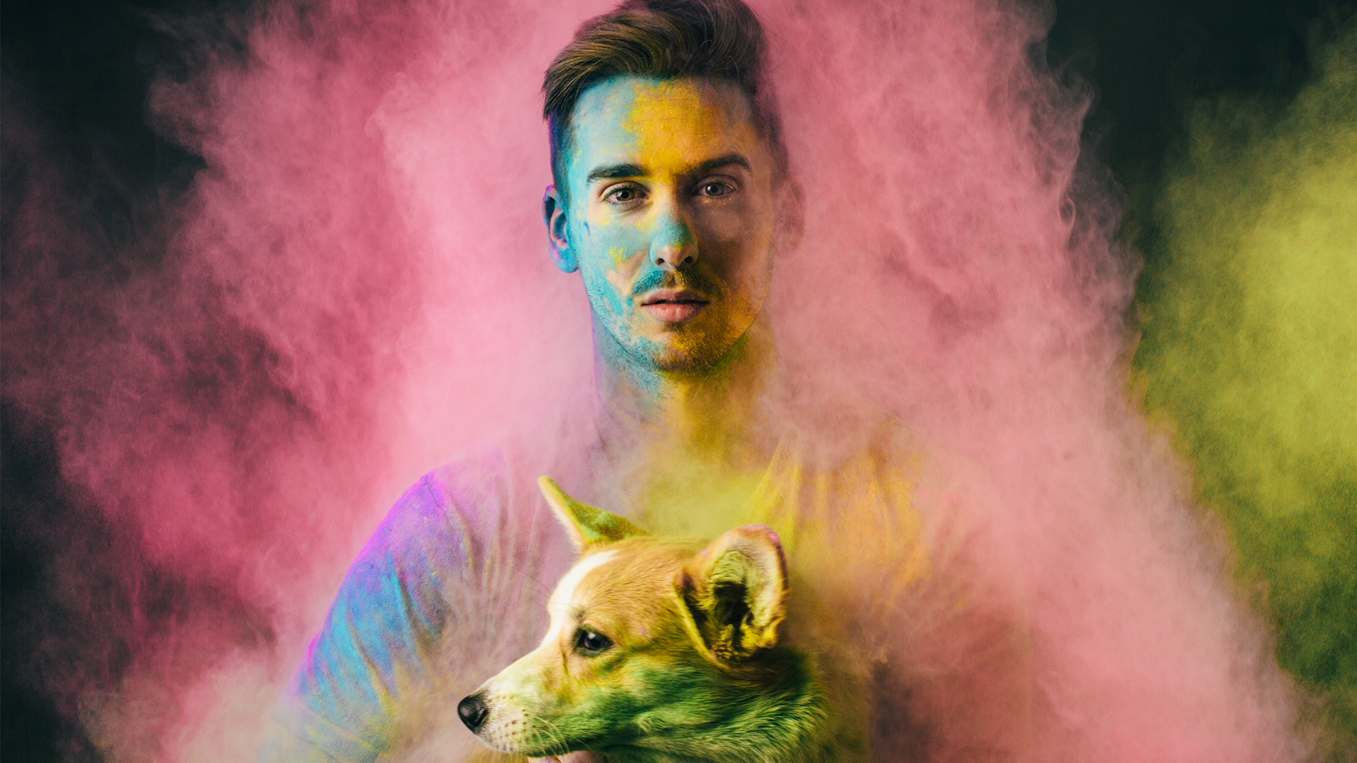 Portrait of Jeff Hodges holding a corgi, both dusted in pink and yellow powder against a dark studio background
