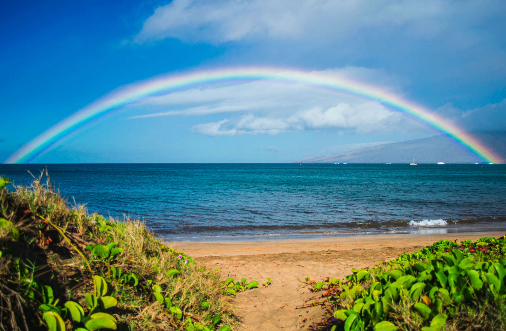 Wide rainbow arching over a beach and blue water after rain